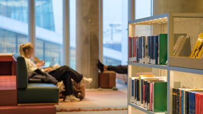 Rader med bøker i hylle. Person sitter og leser i en sofa i bakgrunnen.