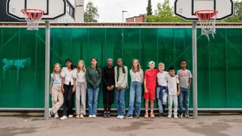 11 skoleelever står ved siden av hverandre mellom to basketballmål i en skolegård.