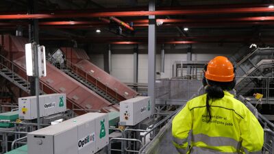 Ryggen på en kvinne med oransje hjelm og gul arbeidsdress på en bro i et sorteringsanlegg. På ryggen hennes står det: "Sammen gjør vi Oslos viktigste jobb!"