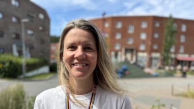 Smilende kvinne i hvit t-skjorte og pride-bånd foran bygg i sol.