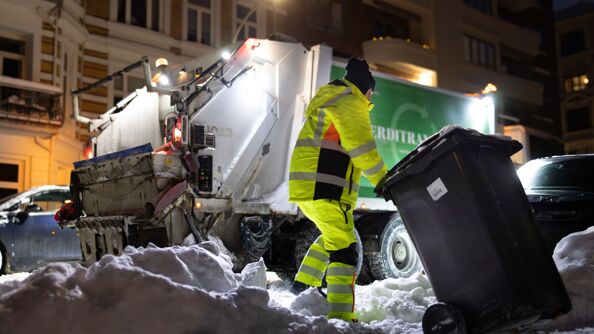 En renovasjonsarbeider i gul refleksvest og jakke drar en søppelkonteiner gjennom en snødekt gate. Bakgrunnen viser en renovasjonsbil og parkert biler, mens snøen ligger tungt i bymiljøet. Scenen foregår om kvelden med lys fra bilen.