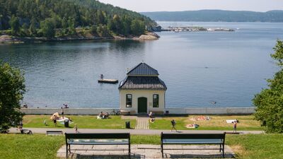 Utsikt over et badeområde ved fjorden på en sommerdag. I forgrunnen står to benker på en gressbakke.