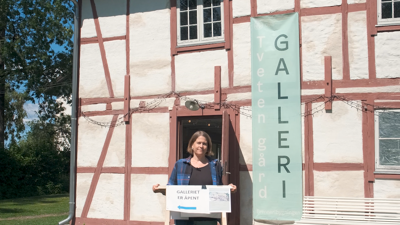 Person står på en gressplen foran et historisk bindingsverkshus med banneret «Tveten gård galleri». Personen holder et skilt med teksten «Galleriet er åpent». Bygningen har hvite vegger, rødt bindingsverk og teglsteinstak, og området er omgitt av grøntarealer.