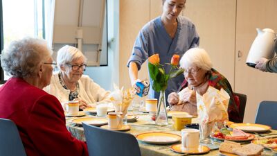 Eldre kvinner sitter rundt er bord og spiser frokost. En pleier i uniform heller i et glass.