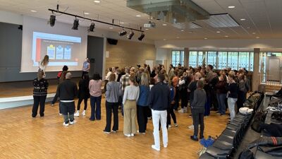 Bildet viser en gymsal hvor to elever, to unge jenter, står på en scene og en stor gruppe med foreldre står forran og hører på hva de sier.