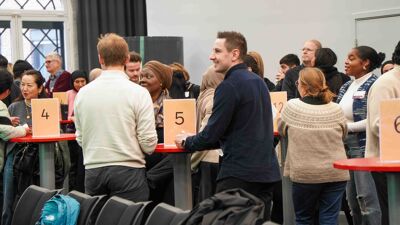 Mange mennesker stående rundt runde bord med nummerlapper på.