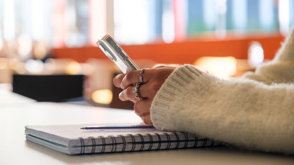 Skoleelev holder en mobiltelefon oppå pulten sin i et klasserom.