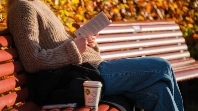 En dame sitter på en benk og leser en bok. det er høstfarger i bakgrunnen.