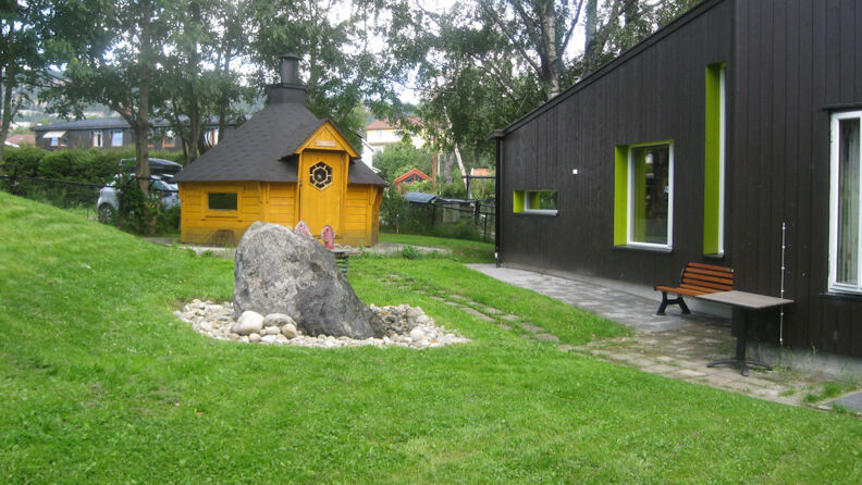 Uteområde med oransje lekehus, benk, gress og en sten, svartmalt barnehage på siden