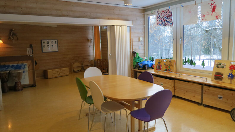 Trefarger rom med stoler, bord, hyller, barnetegninger på veggen 