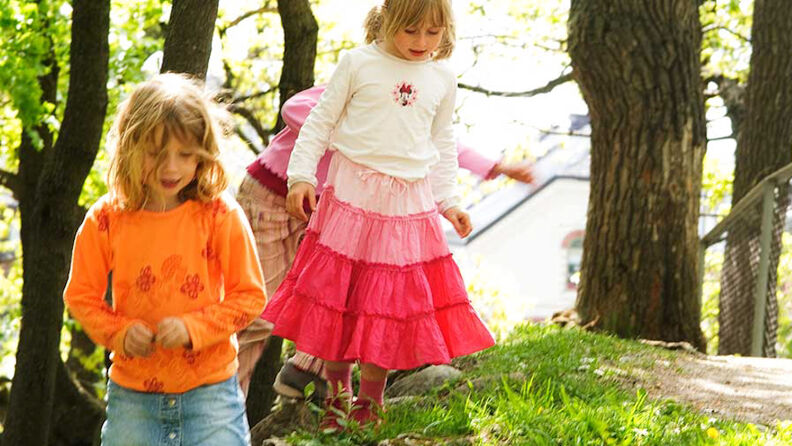 To barn går i gresset i barnehagen, omgitt av trær