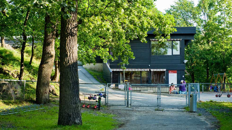 Inngangspartiet til barnehagen, med stålport, huske og barn som leker utfor en svartmalt barnehage omgitt av trær