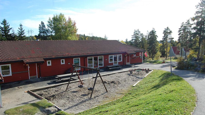 Framsida av rødmalt barnehage med huske som står i sand, 2 benker og gress ovenfor og trær rundt