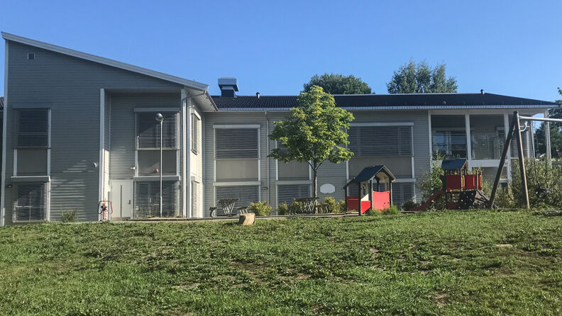 Uteområde med huske og andre lekeapparater, trær og gress, i bakgrunnen gråmalt barnehage