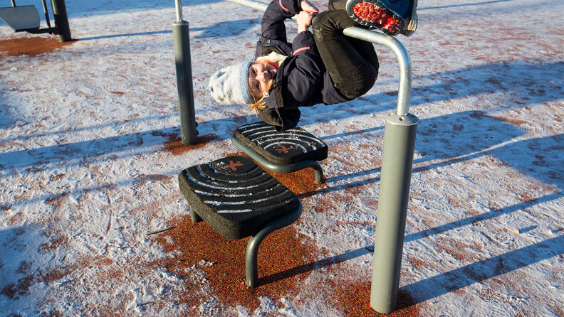 En jente på rundt 6 år i lue og vinterklær henger opp ned i et treningsstativ på en idrettsbane. Det er et tynt snødekke på idrettsbanen.