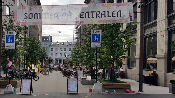 Gåate med trær og benker på begge sider. Banner med påskriften &quot;sommer på sentralen&quot; mellom to stolper. 