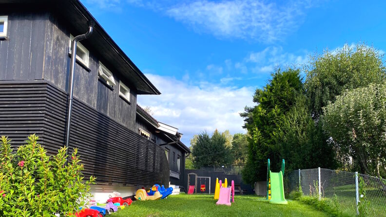 Uteområde med sklie og leker som står på gress,  lekene står  inntil sortmalt vegg til barnehagen, trær på siden og i bakgrunnen