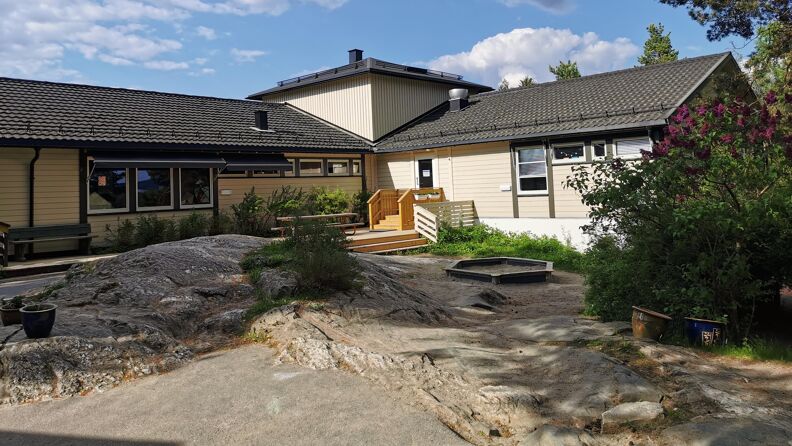 Bilde av barnehagen fra utsiden med benker, sandkasse og grønne busker.