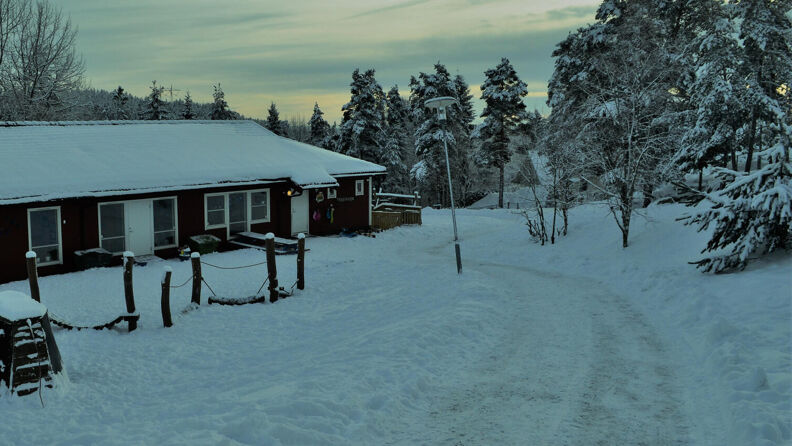 Framsiden av rødmalt barnehage med lekeapparater i snøen og trær rundt