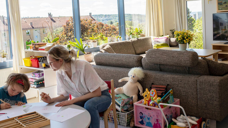 Et barn og en kvinne sitter i et lyst rom ved et barnebord og tegner, med andre leker og sofa og tv i bakgrunnen.