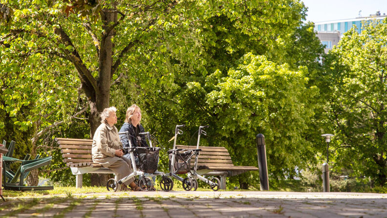 To eldre kvinner sitter ute på en benk med to gåstoler foran seg i en park med grønne trær i bakgrunnen.
