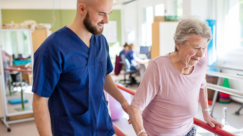 Fysioterapaut hjelper smilende pasient med gåtrening i helsehusets rehabiliteringsrom.