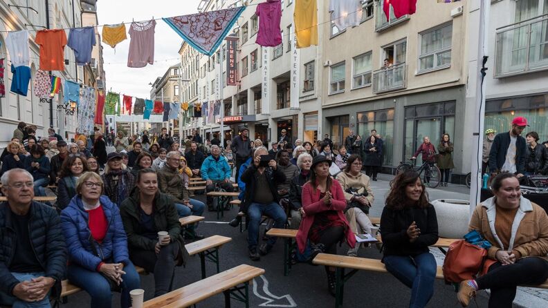 Publikum sitter på penger gjennom gaten mellom bygningen henger det mange klessnorer med klær. 