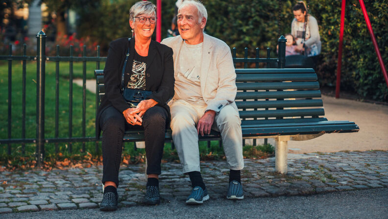 Eldre mann og kvinne sitter i hyggelig dialog på en benk foran en lekeplass i kveldssol.