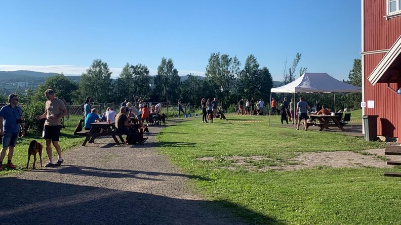 Sommer på gården og mange folk sitter på benker på plenen utenfor det store, røde huset.