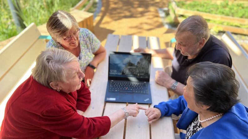 Gjester ved seniorsenteret sitter ute på benker rundt en PC