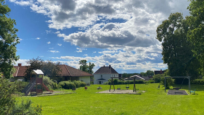 En stor gressplen med lekestativ og to huskesett, omgitt av grønne trær og busker. Det er sol og skyer på himmelen. 