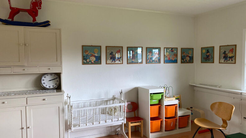Et hvitmalt oppholdsrom med barnlige bilder på veggen, en barneseng og fargerike oppbevaringsbokser i en hylle. På toppen av et skap står en liten, rød gynge-hest. 