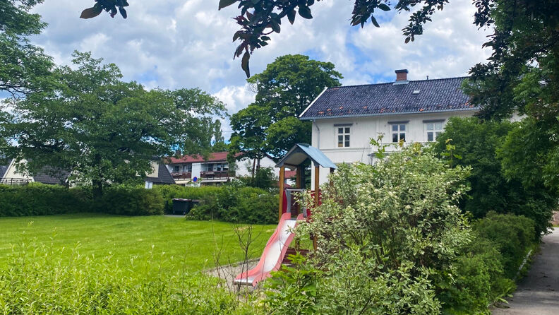 En stor gressplen foran et hvitmalt hus, omgitt av grønne trær og busker. På plenen står et lekestativ med sklie. 