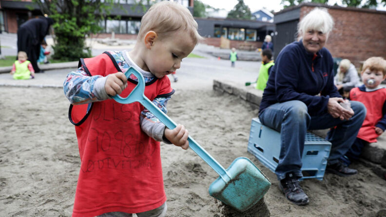 Bilde av et barn i en stor  sandkasse som holder en spade. En dame som jobber i barnehage ser på og smiler.