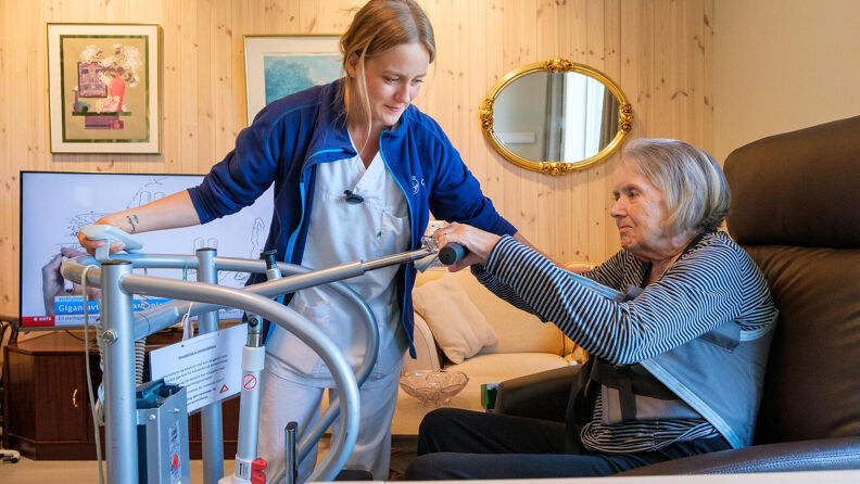 Pleier hjelper beboer opp fra en skinnstol i en tv stue.