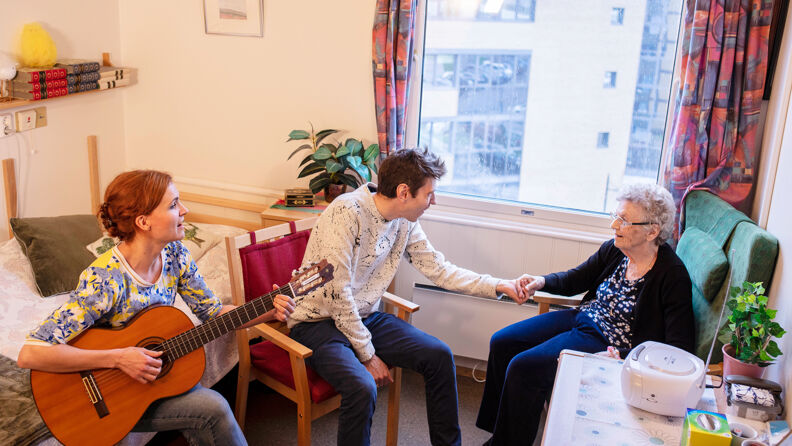 Beboer sitter i rommet sitt med to andre personer hvor den ene spiller gitar.