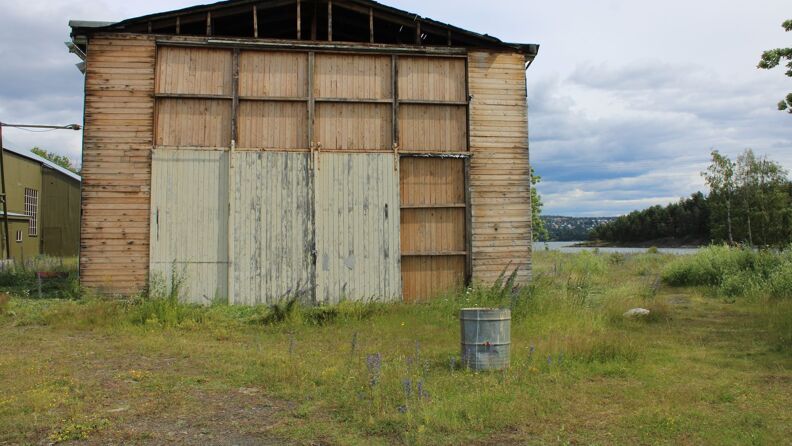 A small old and weathered airplane hangar from the old sea plane airport on Gressholmen, surrounded by long grass and purple flowers. The weather is cloudy with dark clouds in the background