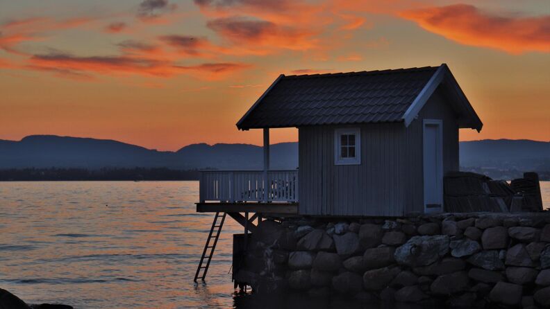 A small bath house on Lindøya right next to the fjord. The weather is nice and there is a sunset making the sky orange.