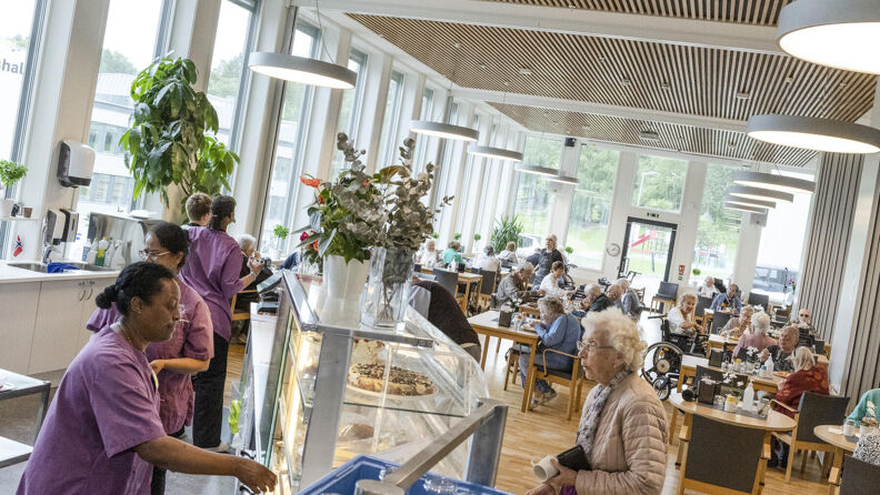 En stor kantine med mange bord og stoler. I kantina er det mange eldre mennesker og ansatte. 