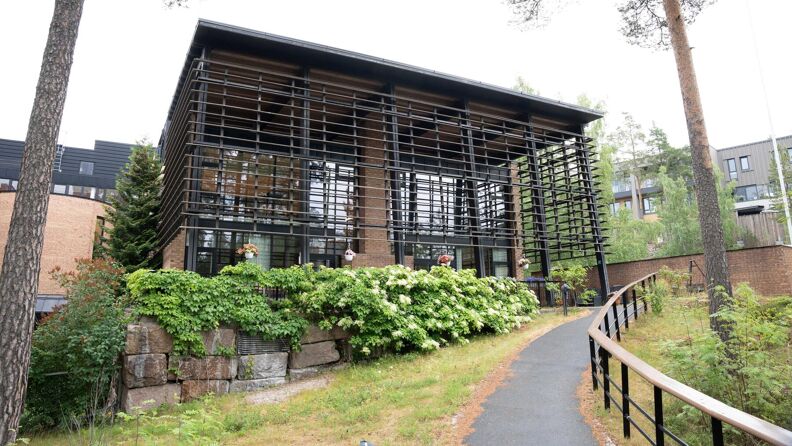 Et stort brunt og svart bygg med store vinduer og ytre riller. Foran bygget er det en gangvei med et gjerde, omringet av blomster, to furutrær og andre vekster.