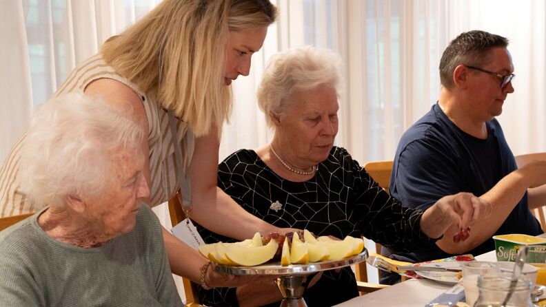 Fire mennesker ved frokostbordet. To beboere blir servert honningmelon av en sykepleier. På bordet ligger det mat av ulike sorter.