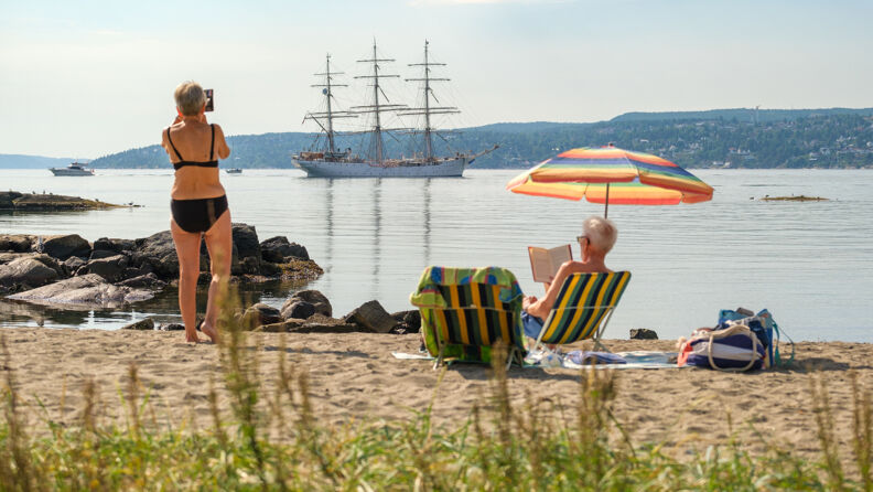 An old couple relaxing at the beach. A man is sitting in a sun chair reading a book and a woman is taking a picture of a large sailboat in the fjord.