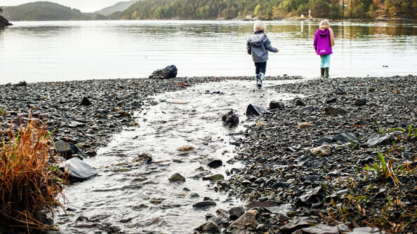 To barn i går langs en strand, med ryggen mot kameraet. I forgrunnen er bakken steinete og en bekk renner gjennom midten av bildet ut i vannet. Trærne i bakgrunnen har høstlige farger.