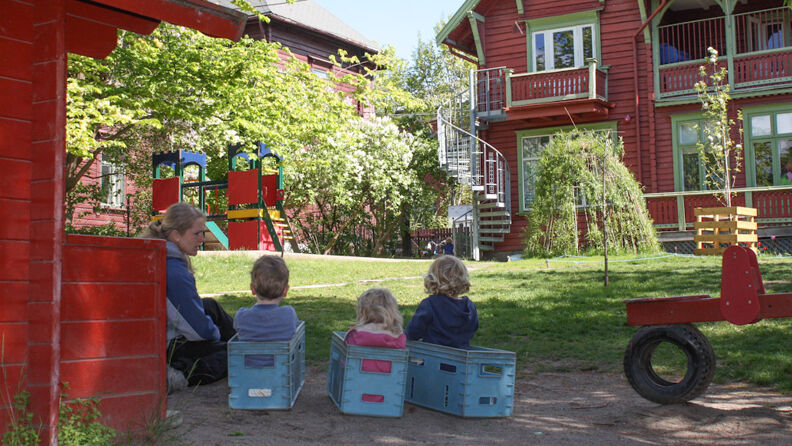 1 voksen og 3 barn sitter med ryggen til og ser mot barnehagens bygning og lekeplass. Det er en solfylt dag med mye grøntområde rundt. 