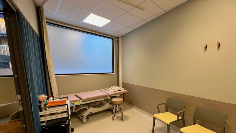 Behandlingsrom med behandlingsbenk, to stoler og et stort vindu som er dekket med folie.