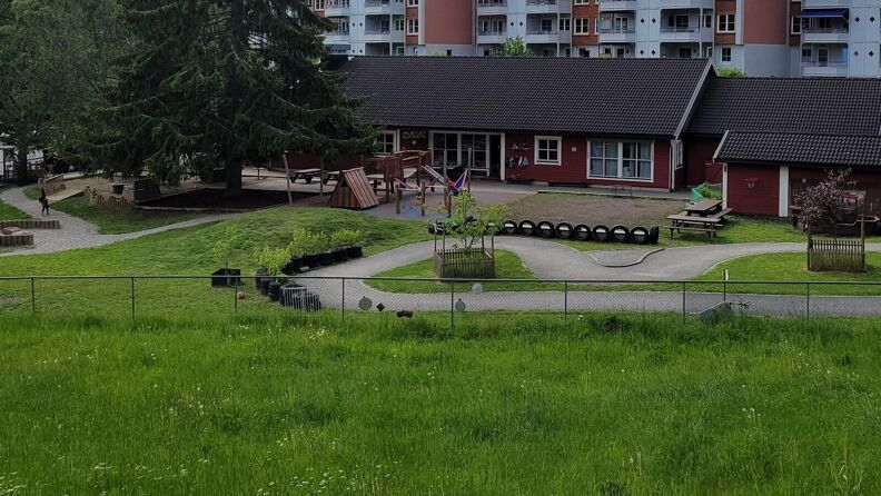 Uteområde med sklie og trapp, huske, benker og en sykkelløype, i bakgrunnen en rødmalt barnehage