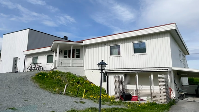 Hvitt trehus med skråtak omgitt av trær og hage. Asfaltert oppkjørsel og parkeringsplass. To hvite biler står parkert foran huset.
