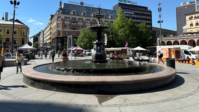 Bilde av fontenen på Youngstorget med vann i. Fint vær og grønne blader på trærne i bakgrunnen. 