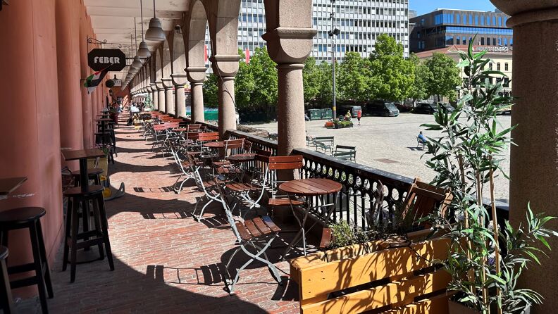 Bilde fra basaren på Youngstorget. Caféstoler og -bord, utsikt ned mot torget. Fint vær og grønne blader på trærne. 