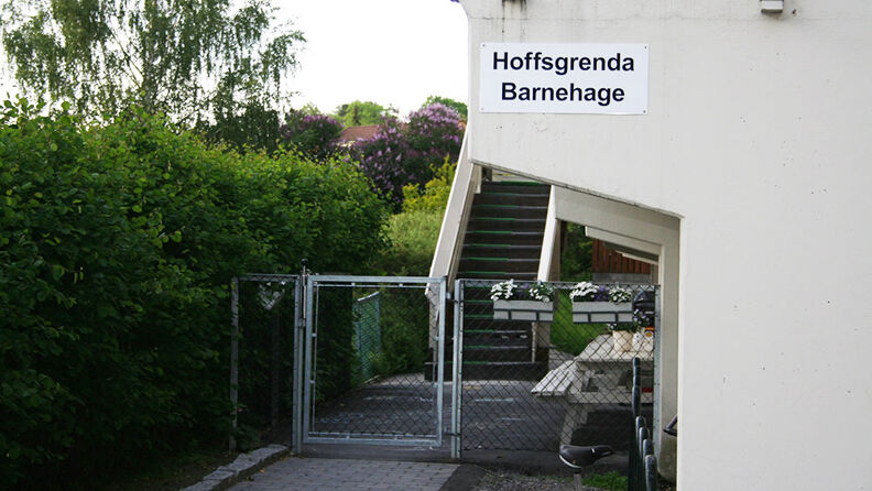 Inngangspartiet til barnehagen, med hekk, stålport og deler av hvitmalt murbygning